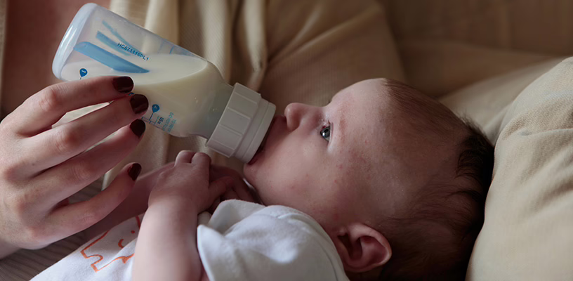 A baby lying in his mom's arms, drinking breast milk from a bottle his mom is holding.