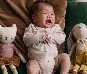 A baby between two stuffed animals, crying because he is gassy.