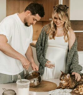 A man making breakfast for his pregnant partner while she strokes their cat.