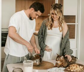 A man making breakfast for his pregnant partner while she strokes their cat.
