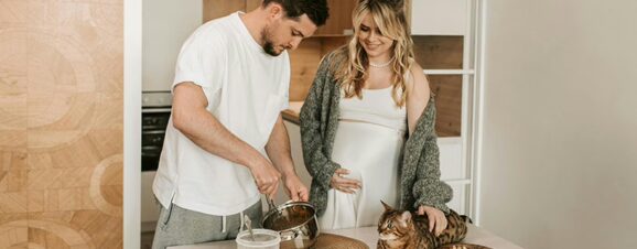 A man making breakfast for his pregnant partner while she strokes their cat.