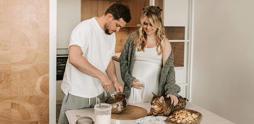 A man making breakfast for his pregnant partner while she strokes their cat.