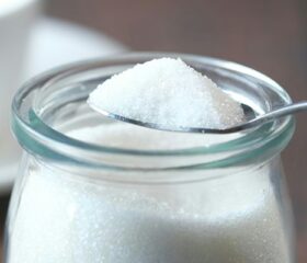 A tablespoon of sugar above a jar of sugar.
