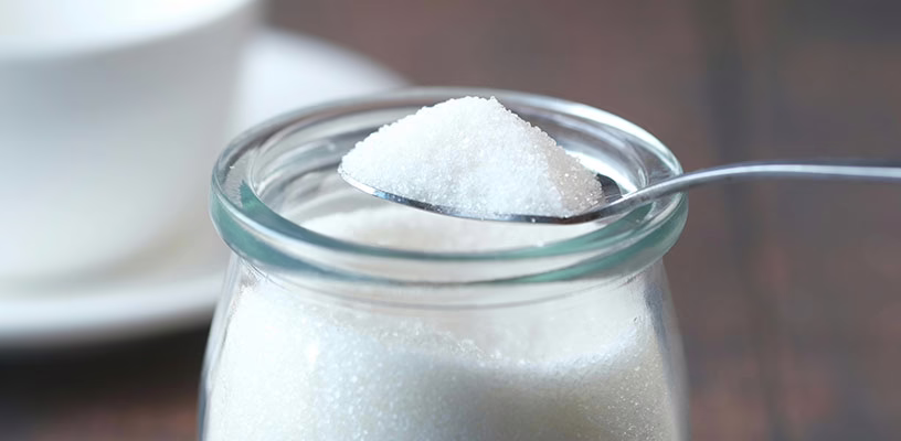 A tablespoon of sugar above a jar of sugar.