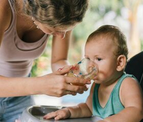A baby sitting in a high chair drinking water from a cup that his mom is holding.
