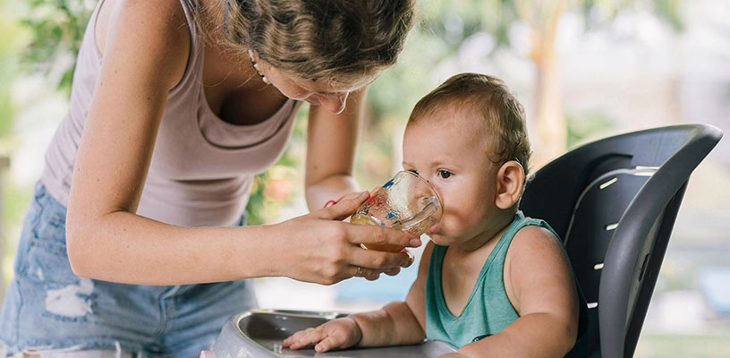 A baby sitting in a high chair drinking water from a cup that his mom is holding.