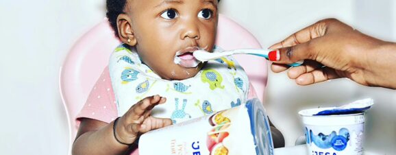 An adult feeding a spoonful of yogurt to a baby sitting in a highchair.