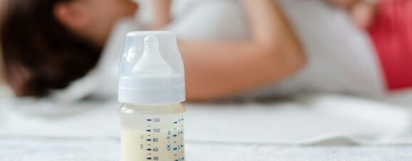 Baby bottle filled with milk in front of a mom playing with her baby.