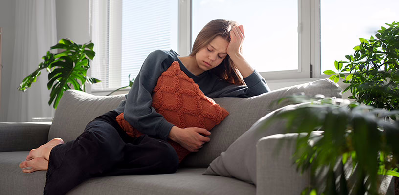 Woman sitting on a sofa suffering from cramps, unable to tell whether they're the result of PMS or pregnancy.