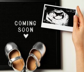 Woman's hand holding an ultrasound image of her baby over a sign that reads "coming soon," with a pair of baby's sandals on it.
