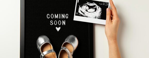 Woman's hand holding an ultrasound image of her baby over a sign that reads "coming soon," with a pair of baby's sandals on it.