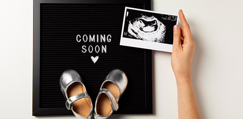 Woman's hand holding an ultrasound image of her baby over a sign that reads "coming soon," with a pair of baby's sandals on it.