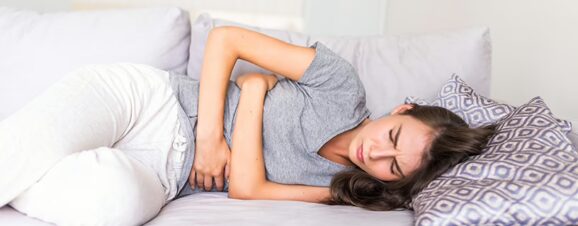 Woman in pain from period cramps, lying on her side on the couch and holding her stomach.