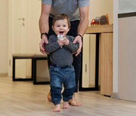 An 12-month-old baby being supported by his father and taking a few steps.