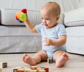 A 7-month-old baby sitting up, smiling, and playing with toys.