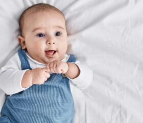 A 7-week-old baby in overalls lying on his back while smiling and laughing.