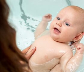 Mother lightly dipping baby in filled bathtub while supporting his head above the water.