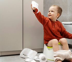 Toddler sitting on the potty, holding up a ply of toilet paper with a roll tossed on the floor next to him.