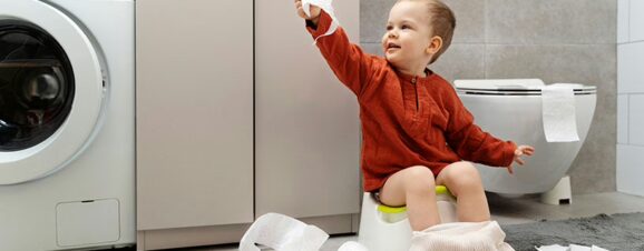 Toddler sitting on the potty, holding up a ply of toilet paper with a roll tossed on the floor next to him.