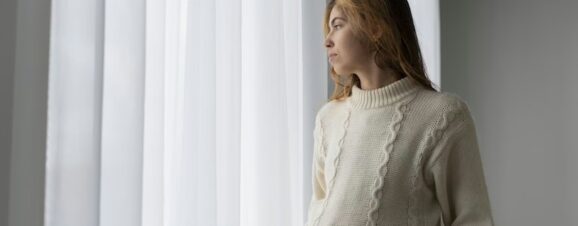 Pregnant woman staring sadly through her curtains.