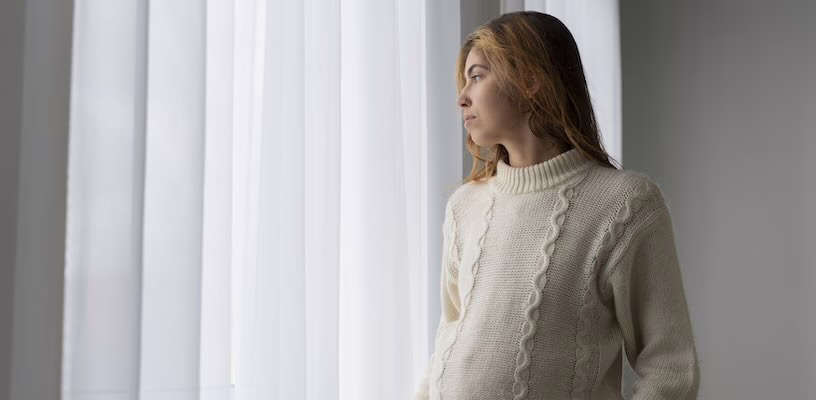 Pregnant woman staring sadly through her curtains.